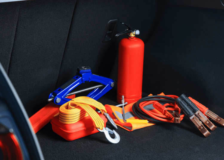 collection of emergency items in trunk of car: red triangle, red fire extinguisher, jumper cables, gloves, and more.